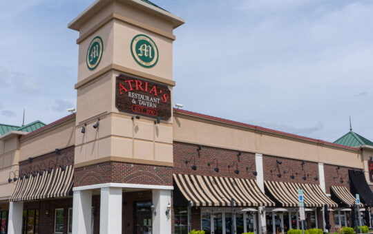 A corner view of Atria’s Restaurant & Tavern, featuring a tall clock tower, striped awnings, patio seating, and potted plants outside the brick building under a partly cloudy sky.