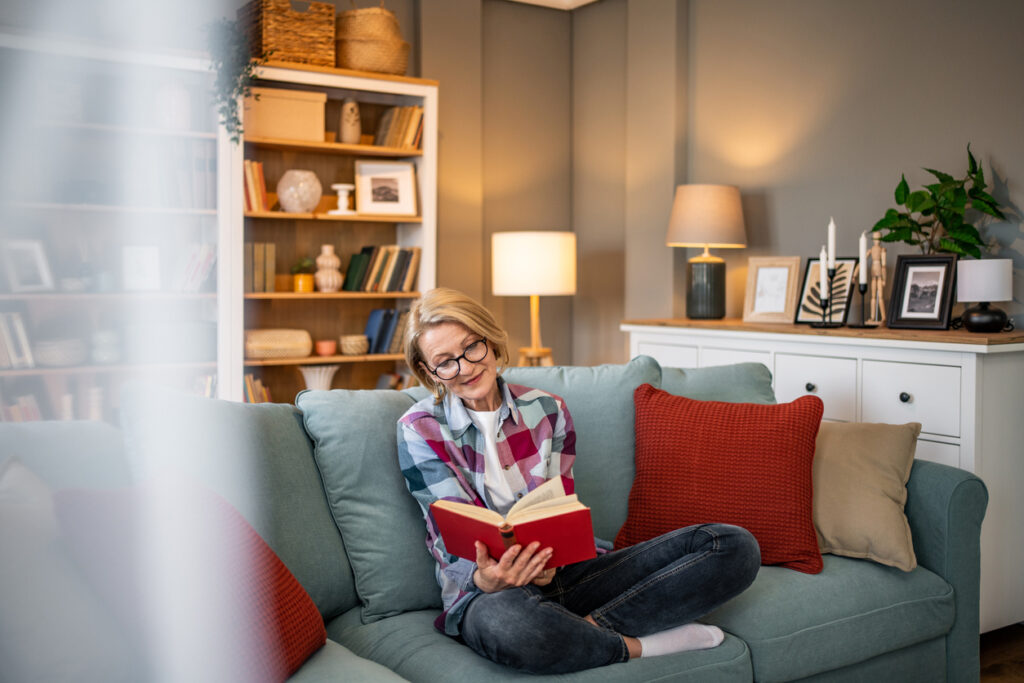 One mature woman, happy mature woman sitting on a sofa in her home reading a book
