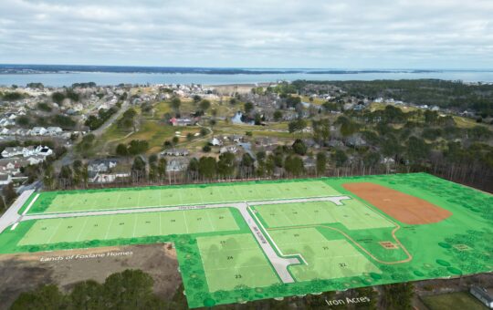 Aerial view of a residential area showing a planned housing development lot, marked in green, next to a baseball field and surrounded by trees, with houses and a body of water in the background.