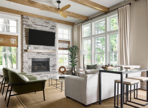 Modern living room with large windows, beige sofa, green armchairs, stone fireplace, TV mounted above, wooden ceiling beams, indoor plant, and a dining table with stools on a woven rug. Natural light fills the space.