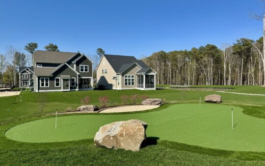 A backyard with a putting green, sand bunker, and large rocks in the foreground; two modern houses and a wooded area are visible in the background under a clear blue sky.