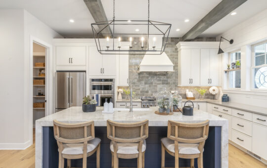 Modern kitchen with white cabinets, a marble island with three wicker barstools, stainless steel appliances, stone backsplash, pendant light fixture, and decorative plants. Large window lets in natural light.