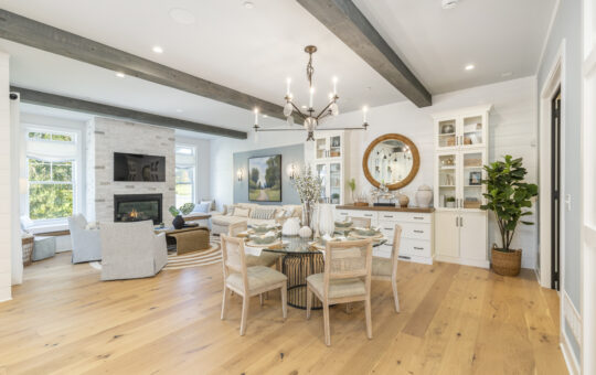 Bright open-concept living and dining room with light wood floors, neutral furnishings, a dining table set for six, a fireplace, and large windows letting in natural light. Exposed ceiling beams and modern decor accents throughout.