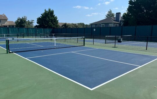 A sunny outdoor pickleball court with blue and green surfaces, surrounded by a black chain-link fence and green privacy screens. Trees and buildings are visible in the background.