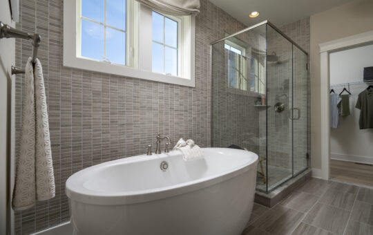 Modern bathroom with a freestanding white bathtub, folded towel on top, glass-enclosed shower, gray tile walls and floor, large window, and a towel hanging on a rack. Closet is visible through an open doorway.