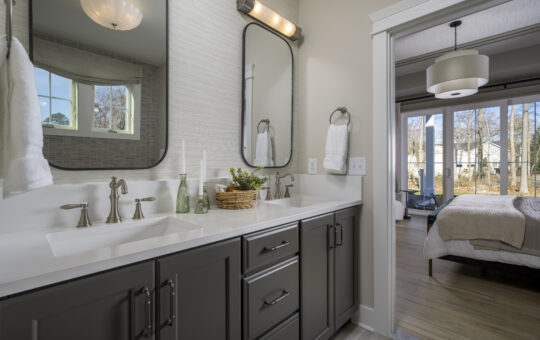Modern bathroom with double sinks, dark cabinets, two tall mirrors, wall-mounted lights, and towel rings with white towels. The bathroom connects to a bright bedroom with large windows and a view outside.