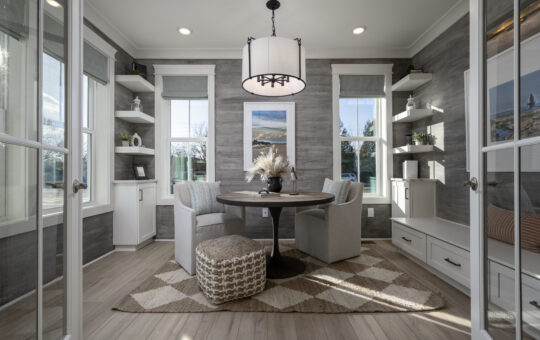 A modern home office with two armchairs around a round table, a patterned rug, built-in shelves, and large windows letting in natural light. A pouf sits on the rug, and a chandelier hangs above the table.