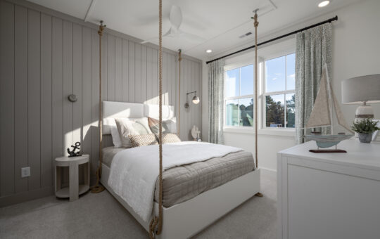 A bright, nautical-themed bedroom featuring a bed suspended by thick ropes, light gray paneled walls, a round nightstand with an anchor decor, a model sailboat, and large windows with patterned curtains.