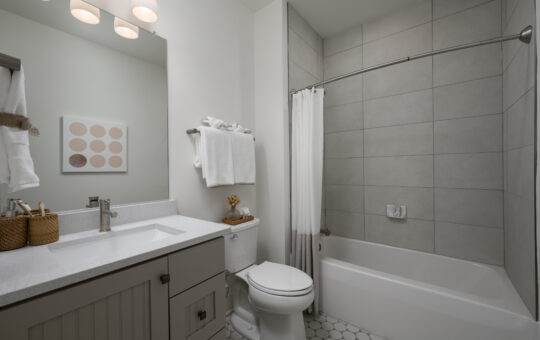 Modern bathroom with a white countertop, sink, and toilet, white hexagonal floor tiles, a bathtub with a white shower curtain, gray tiled walls, and towels neatly folded on a rack above the toilet.