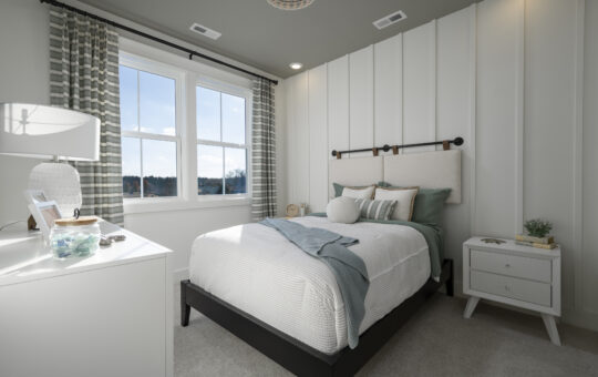 A modern bedroom with a neatly made bed, green and white bedding, a white nightstand, lamp, and a dresser. Large windows with curtains let in natural light. The walls are white with vertical paneling.