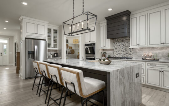 Modern kitchen with a large marble island, four cushioned bar stools, stainless steel appliances, white cabinets, and pendant lighting. The backsplash features a mosaic design behind a stove with a dark range hood.