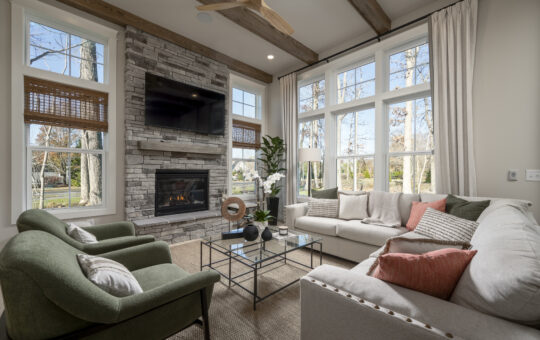 A cozy living room with large windows, a stone fireplace with a mounted TV, two green armchairs, a beige sectional sofa with pink pillows, a glass coffee table, and indoor plants. Sunlight streams through the windows.