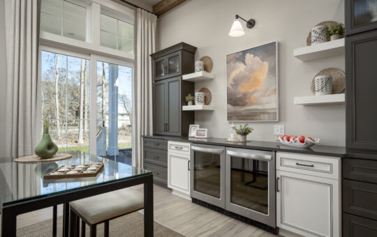 A modern kitchen with large windows, dark cabinets, white drawers, two wine coolers, open shelves decorated with vases and baskets, and a framed painting of clouds on the wall above the counter.