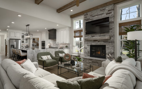 A modern open-concept living room and kitchen with a stone fireplace, wall-mounted TV, sectional sofa, green armchairs, and a kitchen island, featuring wood accents, large windows, and natural light.