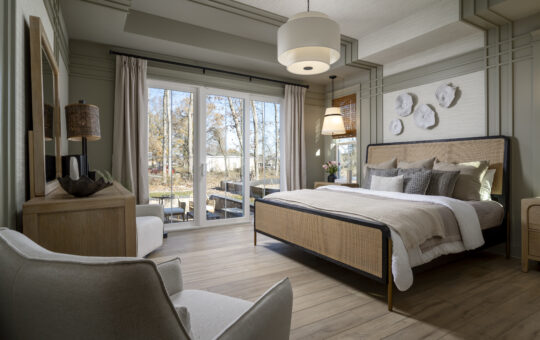 A modern bedroom with a wooden floor, a large bed with neutral bedding, a soft armchair, and floor-to-ceiling windows letting in natural light. Wall art, lamps, and outdoor trees are visible through the windows.