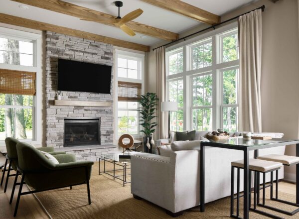 Modern living room with large windows, beige curtains, stone fireplace, wall-mounted TV, light sofa, two green armchairs, a tall plant, and a small dining table with stools on a neutral rug. Natural light fills the space.