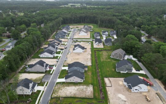 Aerial view of a suburban neighborhood with newly built houses, some empty lots, green lawns, and surrounding trees. Roads separate rows of homes, and construction materials are visible on several properties.