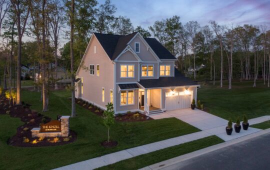 Two-story modern house with large windows and a front porch, surrounded by grass and trees at dusk. A lit sign reading THE KNOLL is visible in the landscaped yard near the street.