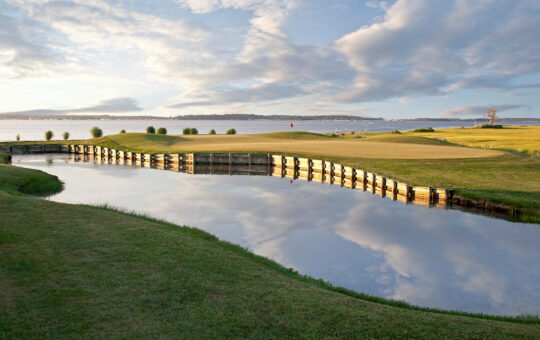 A scenic golf course with a green bordered by wooden barriers next to a calm water hazard, reflecting the sky and clouds. In the background, there is a lake and open grassy areas under a partly cloudy sky.