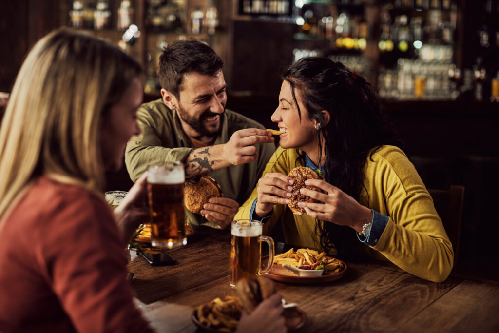 Happy man feeding his girlfriend with French fries while eating hamburgers in a pub. 