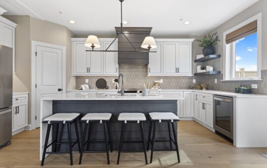 Modern kitchen with white cabinets, a large island with four stools, stainless steel appliances, two pendant lights, and a window letting in natural light. Light wood flooring and neutral tones create a clean, bright space.