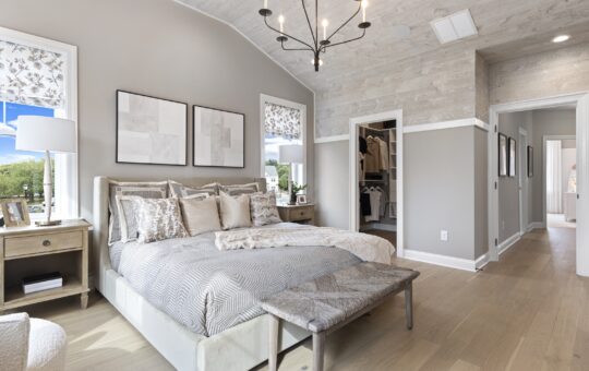 A modern bedroom with a large bed, neutral bedding, two abstract art pieces above the headboard, nightstands with lamps, a bench at the foot of the bed, wood floors, and a walk-in closet visible through an open door.
