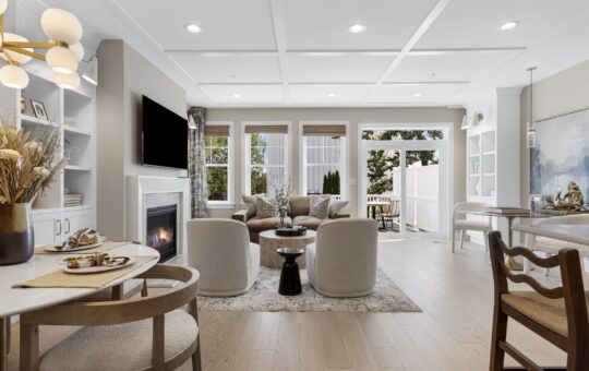 Bright, modern living and dining area with neutral tones, round table set for two, cozy seating around a fireplace and TV, large windows, light wood floors, and sliding doors opening to a sunny patio and fenced backyard.