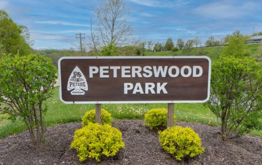 A wooden sign reading Peterswood Park stands amid green shrubs and mulch, with a grassy field, trees, and a partly cloudy sky in the background.
