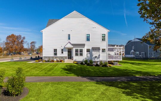 A white two-story house with a small porch sits on a neatly mowed green lawn under a clear blue sky. Decorative plants and shrubs border the front, and neighboring houses are visible in the background.