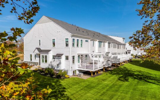 A row of modern white townhouses with large windows and back decks overlooks a well-maintained green lawn, framed by leafy trees under a clear blue sky.