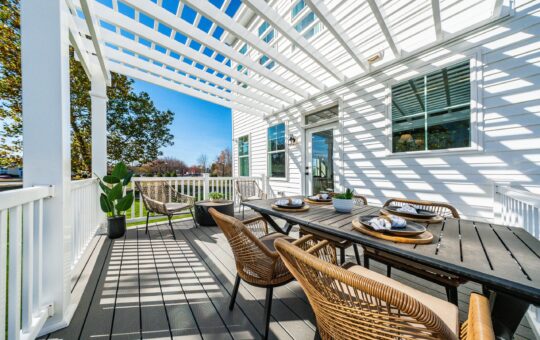 A modern outdoor patio with a pergola features a dining table set for four, wicker chairs, potted plants, and sunlight casting striped shadows across the deck. White railings and a green lawn are visible in the background.