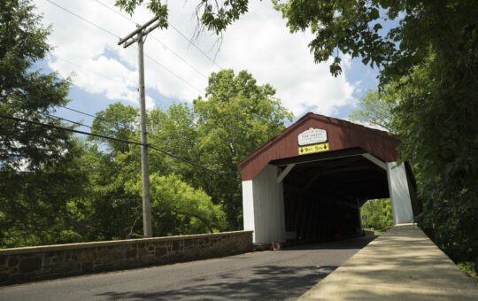 A red and white covered bridge spans a road surrounded by green trees under a partly cloudy sky. A stone wall lines the left side of the road, and a yellow clearance sign is visible on the bridge.