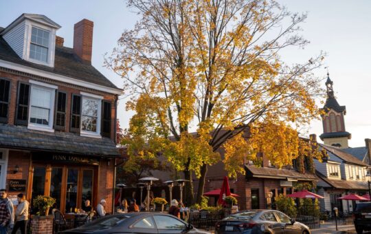 A street scene in autumn shows people dining outdoors by a restaurant with yellow-leaved trees, brick buildings, and parked cars under a clear sky at sunset.