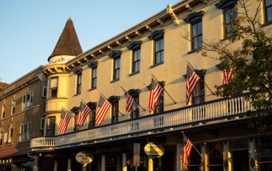 A historic yellow building with a round turret and balcony is decorated with multiple American flags. The warm sunlight casts shadows, highlighting the architectural details and nearby storefronts.