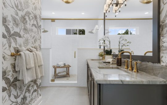 Elegant bathroom with floral wallpaper, double sinks with gold faucets, a large mirror, folded towels on a gold rack, and a spacious glass-enclosed shower with a wooden bench and natural light from high windows.