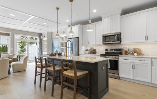 Modern kitchen with white cabinets, stainless steel appliances, a black island with a white countertop, three wooden bar stools, pendant lights, and an open view to the living area and backyard.