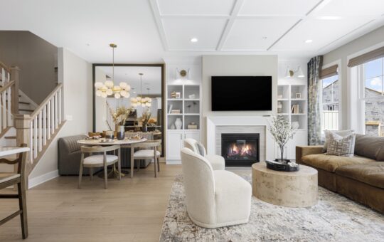 A modern living room with a beige couch, round coffee table, armchairs, and a fireplace under a wall-mounted TV. The dining area and a staircase are visible, with large windows letting in natural light.