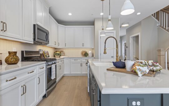 Modern kitchen with white cabinets, stainless steel appliances, a center island with a sink, pendant lights, wood flooring, and decorative accents on countertops. Staircase and hallway visible in the background.