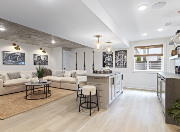 Modern living room with a beige sectional sofa, round coffee tables, wall art, and a kitchen island with stools. Pendant lights hang from the ceiling, and large windows let in natural light. Light wood flooring throughout.