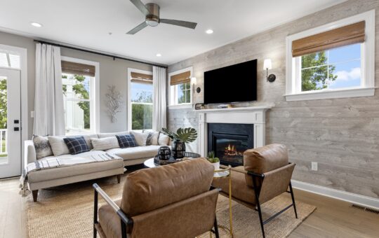 Modern living room with a sectional sofa, two brown leather chairs, a woven rug, a fireplace with a TV above it, large windows with blinds, and neutral decor in natural light.