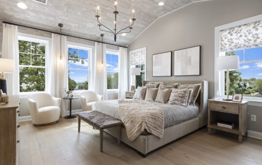 A modern, cozy bedroom with a large bed, neutral bedding, two armchairs by bright windows, wooden nightstands with lamps, light wood floors, and abstract art on the walls. Trees and blue sky are visible outside.
