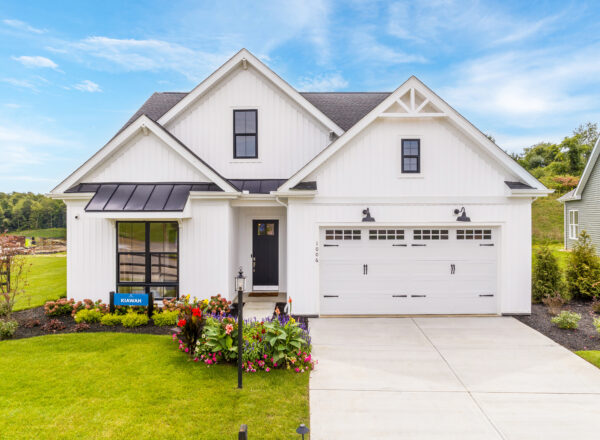 A modern white house with black roof accents, a two-car garage, manicured lawn, and colorful flower beds in front, set against a bright blue sky with scattered clouds.