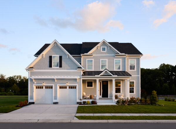 A two-story suburban house with gray siding, white trim, a double garage, and a covered front porch. The home sits on a well-manicured lawn with trees in the background under a blue sky with light clouds.