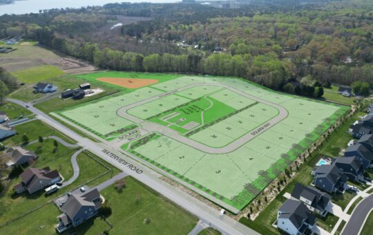 Aerial view of a residential neighborhood with an overlay plan showing a future housing development, numbered lots, streets, open green spaces, and a baseball field, surrounded by trees and existing homes.