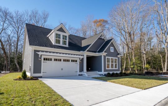 A modern gray house with white trim, a double garage, and a large driveway, surrounded by green lawn and trees with bare branches under a clear blue sky.