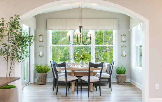 A bright dining area with large windows, a round wooden table set for a meal, six cushioned chairs, green plants in baskets reminiscent of a low-maintenance garden, and a modern black chandelier hanging above the table.