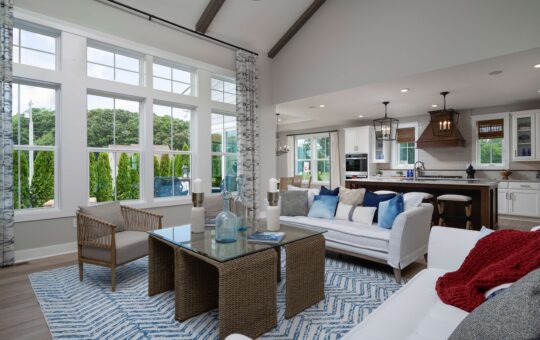 Bright, modern open-concept living room and kitchen with large windows, white sofas, blue accents, wicker coffee table, and patterned rug; greenery visible outside.