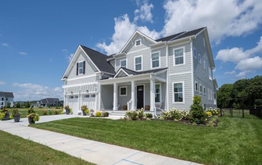 A modern two-story gray house with white trim, a covered front porch, and a well-kept lawn on a sunny day with a blue sky and scattered clouds. There are flower beds and a sidewalk in front.