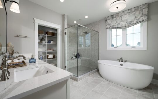 Modern bathroom with a freestanding bathtub, glass-enclosed shower, white vanity with marble countertop, closet shelves with baskets and towels, and a large window with a patterned valance.