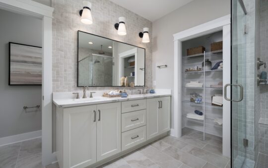 Modern bathroom with a double-sink vanity, large mirror, three lights above, glass shower on the right, and an open linen closet with neatly organized towels and baskets. Light gray and white color scheme.
